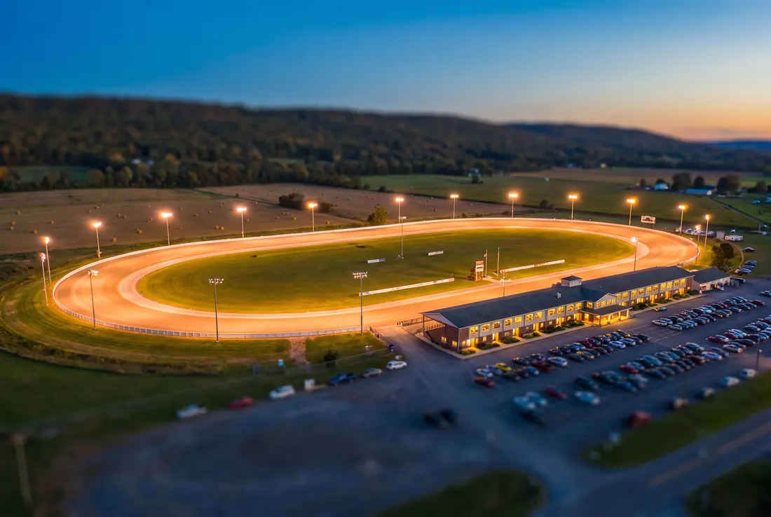 Tilt-shift miniature of a floodlit harness racing oval at dusk with a long grandstand and farmland beyond
