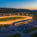 Tilt-shift miniature of a floodlit harness racing oval at dusk with a long grandstand and farmland beyond